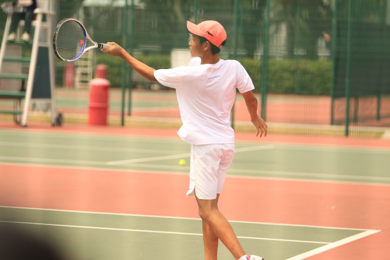 An ACS(I) tennis player tosses the ball up during a serve on an outdoor court under a bright sky.